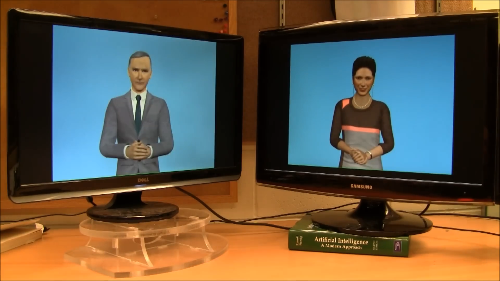 The original 2011 desk: Alan on the left monitor and Sruthi on the right, with blue screen backgrounds, cables, a clear circular support, and an artificial intelligence textbook.